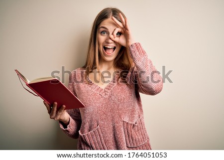 Young beautiful redhead student woman reading book over isolatated white background with happy face smiling doing ok sign with hand on eye looking through fingers