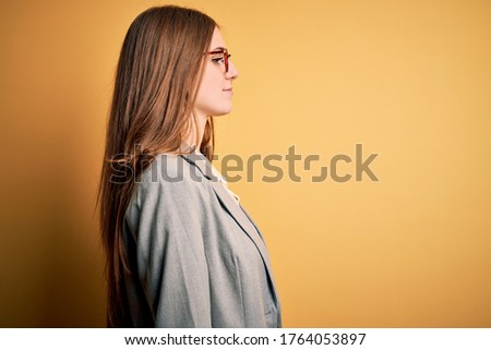 Young beautiful redhead woman wearing jacket and glasses over isolated yellow background looking to side, relax profile pose with natural face with confident smile.
