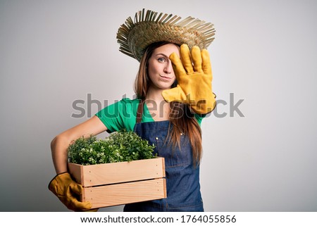 Young beautiful redhead farmer woman wearing apron and hat holding box with plants with open hand doing stop sign with serious and confident expression, defense gesture
