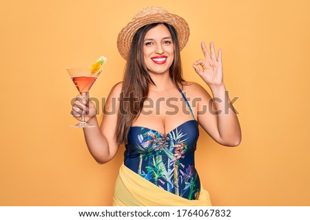 Young hispanic woman wearing summer swimsuit drinking a cocktail over yellow background doing ok sign with fingers, excellent symbol