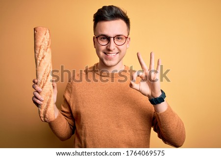 Young handsome caucasian man wearing glasses holding a fresh baguette of bread doing ok sign with fingers, excellent symbol