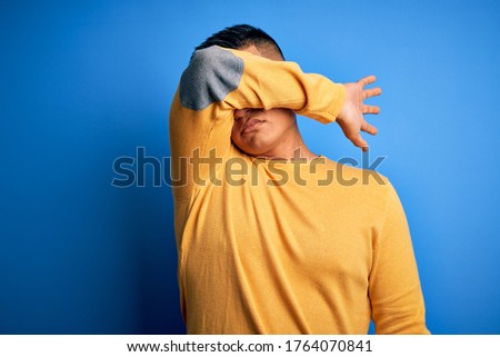 Young handsome latin man wearing yellow casual sweater over isolated blue background covering eyes with arm, looking serious and sad. Sightless, hiding and rejection concept