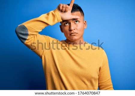 Young handsome latin man wearing yellow casual sweater over isolated blue background making fun of people with fingers on forehead doing loser gesture mocking and insulting.