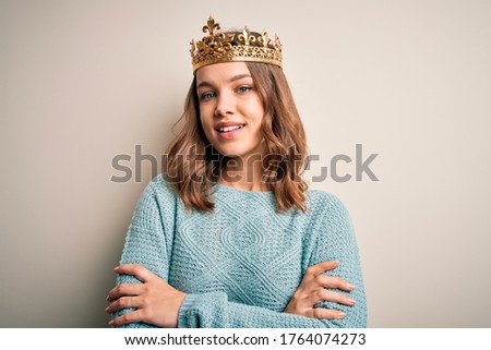 Young blonde girl wearing queen golden crown over isolated background happy face smiling with crossed arms looking at the camera. Positive person.