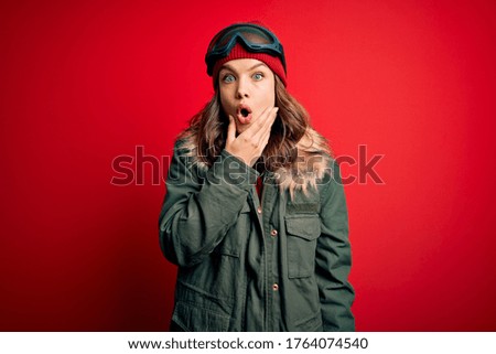 Young blonde girl wearing ski glasses and winter coat for ski weather over red background Looking fascinated with disbelief, surprise and amazed expression with hands on chin