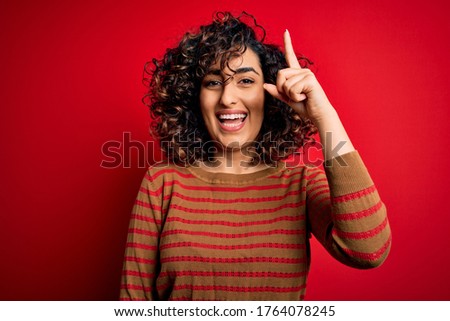 Young beautiful curly arab woman wearing casual striped sweater standing over red background pointing finger up with successful idea. Exited and happy. Number one.