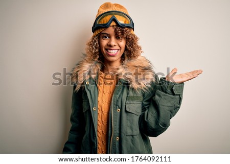 Young african american skier woman with curly hair wearing snow sportswear and ski goggles smiling cheerful presenting and pointing with palm of hand looking at the camera.