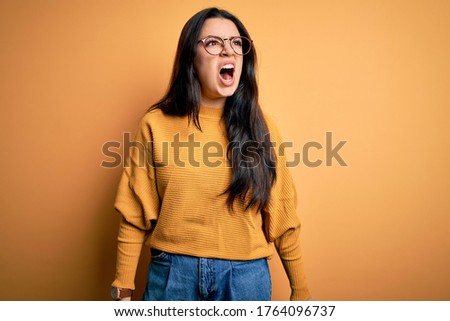 Young brunette woman wearing glasses and casual sweater over yellow isolated background angry and mad screaming frustrated and furious, shouting with anger. Rage and aggressive concept.