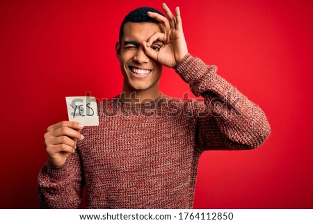 Young handsome african american man holding reminder paper with yes message with happy face smiling doing ok sign with hand on eye looking through fingers