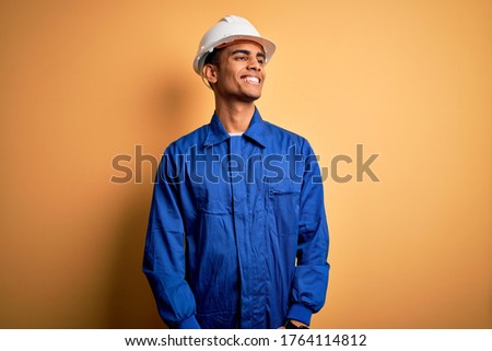 Young handsome african american worker man wearing blue uniform and security helmet looking away to side with smile on face, natural expression. Laughing confident. Royalty-Free Stock Photo #1764114812