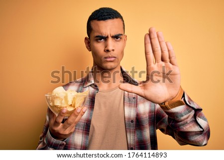 Young handsome african american man holding bowl of chips potatoes over yellow background with open hand doing stop sign with serious and confident expression, defense gesture