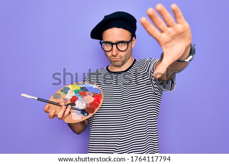 Young artist man with blue eyes wearing french beret and glasses drawing using paintbrush with open hand doing stop sign with serious and confident expression, defense gesture
