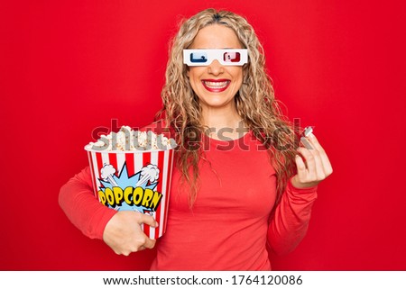 Young beautiful blonde woman watching movie using 3d glasses eating popcorn looking positive and happy standing and smiling with a confident smile showing teeth