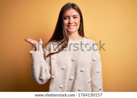 Young beautiful woman with blue eyes wearing casual sweater standing over yellow background smiling cheerful presenting and pointing with palm of hand looking at the camera.