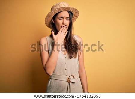 Young beautiful brunette woman on vacation wearing casual dress and hat bored yawning tired covering mouth with hand. Restless and sleepiness.