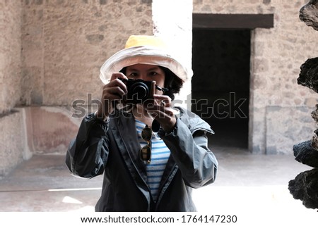 A middle aged Asian woman holding a compact camera close to her face in a ruined site of Teotihuacán Municipality. Dramatic lighting.