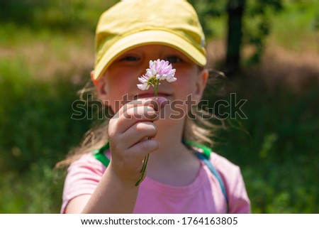 Positive emotional young lady smiles, looks on wild flower that holds in hand. Cheerful pretty child girl in yellow baseball cap, pink shirt, stands on nature background on sunny day. Happy childhood.
