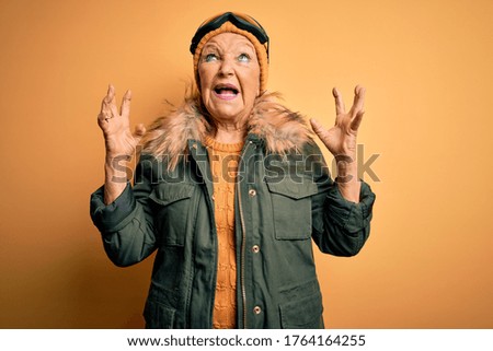Senior beautiful grey-haired skier woman wearing snow sportswear and ski goggles crazy and mad shouting and yelling with aggressive expression and arms raised. Frustration concept.