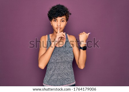 Young beautiful african american afro sportswoman doing exercise wearing sportswear asking to be quiet with finger on lips pointing with hand to the side. Silence and secret concept.