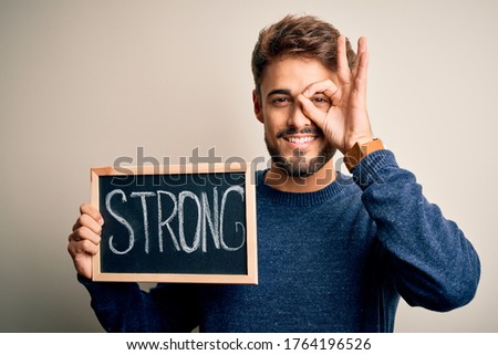 Young man holding blackboard with strong message standing over isolated white background with happy face smiling doing ok sign with hand on eye looking through fingers