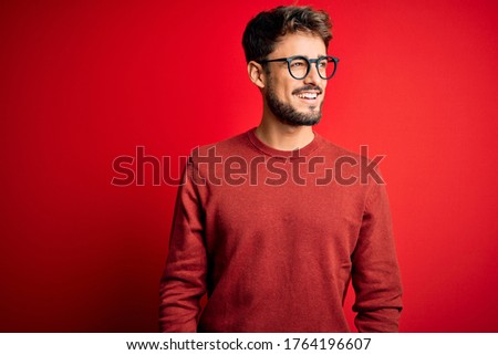 Young handsome man with beard wearing glasses and sweater standing over red background looking away to side with smile on face, natural expression. Laughing confident.