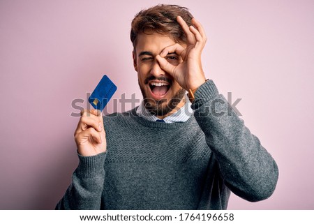 Young customer man with beard holding credit card for payment over pink background with happy face smiling doing ok sign with hand on eye looking through fingers