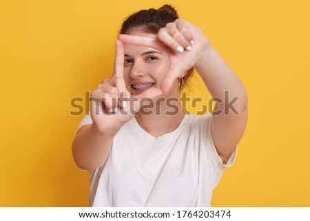 Happy smiling woman making frame in front her face and smiling, young girl having fun posing isolated over yellow background, girl in white casual t shirt with positive expression.