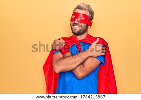 Handsome blond man wearing super hero costume with mask and cape over yellow background hugging oneself happy and positive, smiling confident. Self love and self care