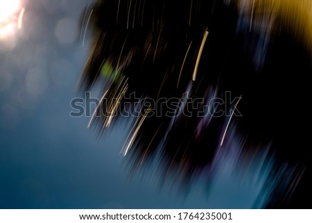 Silhouetted, abstract, motion-blurred, tree, vegetation with golden and multicolor light streaks on blue sky with sun showering off-white hexagonal lens flares