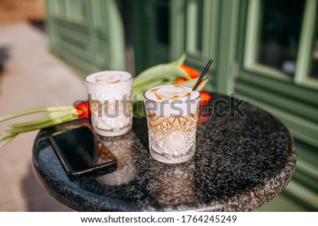 two cups of latte coffee with milk with a picture and with black tubes in glass glasses are on the table near the cafe. On the table are a telephone and a bouquet of flowers