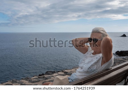 Smiling senior lady sitting on wooden bench with horizon over the sea in background - relaxed people in sea excursion, cheerful retirement concept