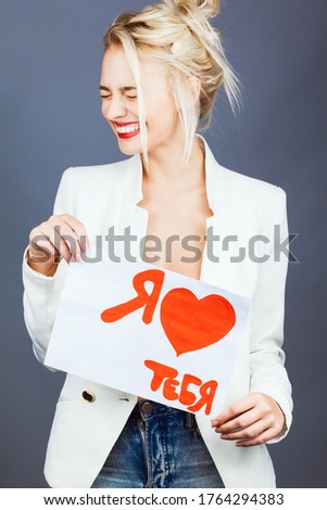Portrait of a young friendly woman in with a beautiful face holds a sheet of paper on grey background. Emotional face. I LOVE YOU