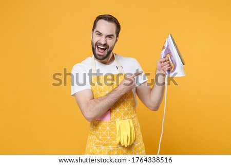 Cheerful crazy young man househusband in apron doing housework isolated on yellow wall background studio portrait. Housekeeping concept. Mock up copy space. Pointing index finger on iron in hand