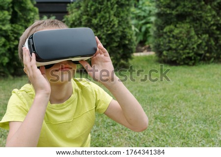 boy in virtual reality glasses on his eyes seat on green grass and watch with maximum attention  something  in virtual reality. Copyspace for text. Impact of new technologies for children