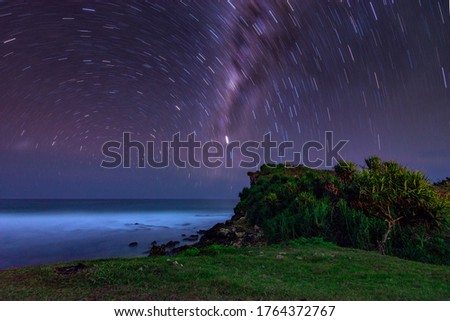 Star trails and Milky Way galaxy on the beach with green hills on the beach of Ngedan, Gunungkidul, Special Region of Yogyakarta.