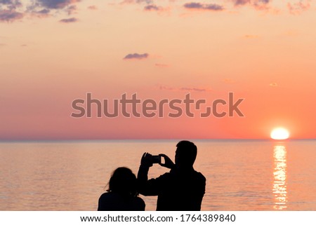 A man photographs a beautiful sunset on a smartphone. Silhouettes of women and men on a blurred background.