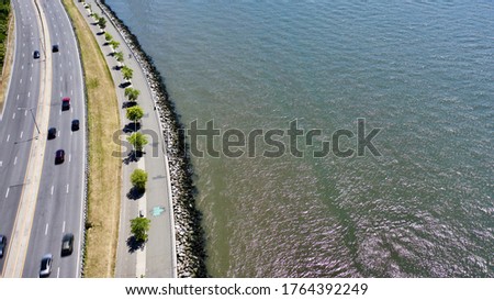 A drone camera view over the Belt Parkway in Brooklyn, New York. The waters of New York Bay is visible on the right of the shot. It is a bright and sunny day with clear blue skies.