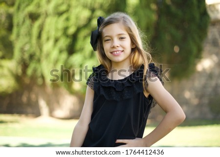Studio shot of cheerful little girl standing outdoors keeps hand on hip, smiles broadly, anticipates for surprise prepared by friends on her birthday.