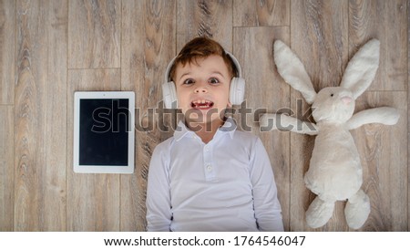 Top view of little kid boy laying on the floor with his rabbit toy and listen to the music in wireless headphones.