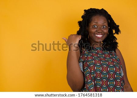 lovely Young afro american with afro hair woman wearing glasses over isolated yellow background pointing aside with forefinger, showing at copy space having news about bargains 