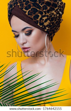 An attractive young woman in a stylish turban made of leopard print fabric on a yellow mango background. Girl with bright makeup. Beach style, palm branch