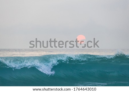 Blue ocean wave in front of beautiful sunset sky with pink sun disk over ocean skyline.