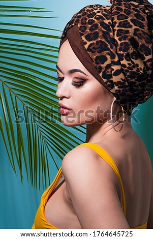 Summer sea and the hot weather, the beach trend. A young beautiful multi-ethnic woman in a leopard turban and yellow swimsuit looks out from behind a palm branch, blue background