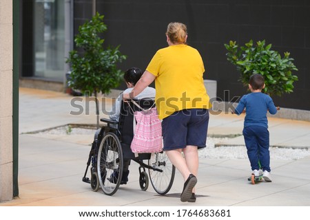 unrecognizable woman pushing a wheelchair with a disabled person, next to a little boy riding a scooter, rear view on a city street
