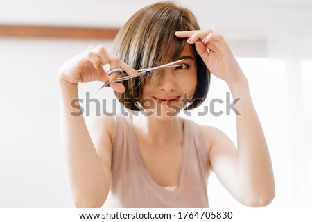 Closeup portrait of happy young Asian woman having her hair cut with scissors at home. She's stay at home during the coronavirus pandemic, Self hair care during quarantine. Royalty-Free Stock Photo #1764705830