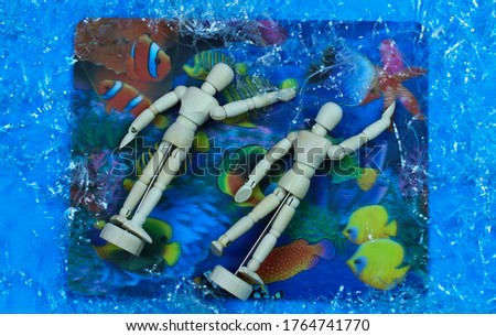 Overhead photo of two wooden mannequins swimming in a sea full of colorful fish.