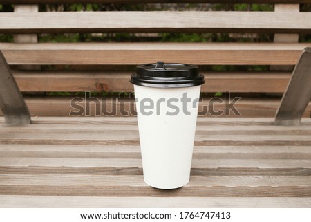 A white cup with a black lid stands on a wooden bench.
