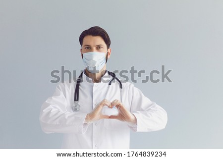 Young male doctor in medical mask showing heart gesture on grey background. Cardiovascular health, charity or donation