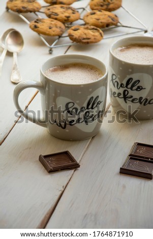 Hot chocolate in two cups, pieces of dark chocolate on a white rustic wood table, freshly baked cookies with chocolate on a baking sheet, two teaspoons, cups hello weekend, top view