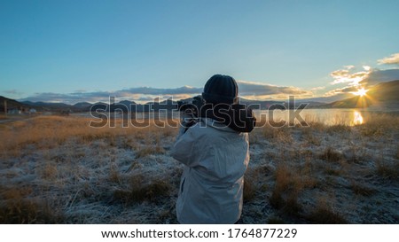 Woman photographer hold camera in warm white winter outfit and bean hat in sunset mountain. Asian female traveler capture sunlight magic moment in winter nature outdoor. tourist vacation trip concept.